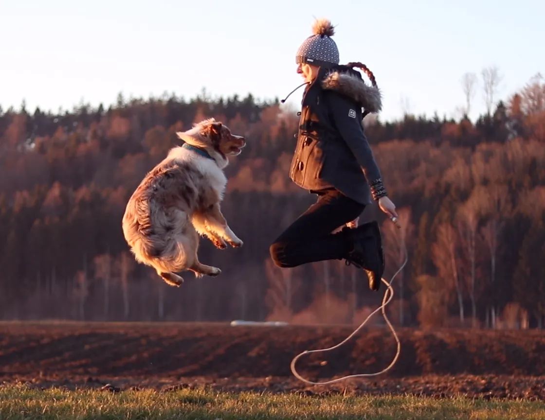 Sophie Paulin springt zusammen mit ihrem Australian Shepherd auf einem Feld in die Luft